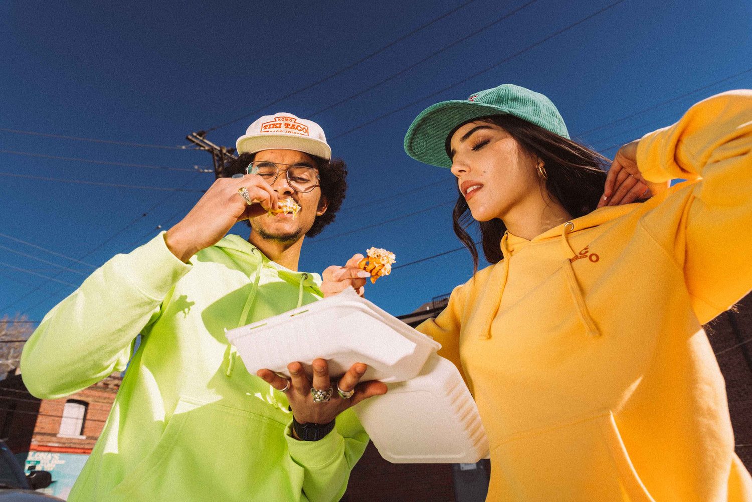 Young guy and girl dressed in Tiki Taco branded merch while enjoying some carne asada fries.
